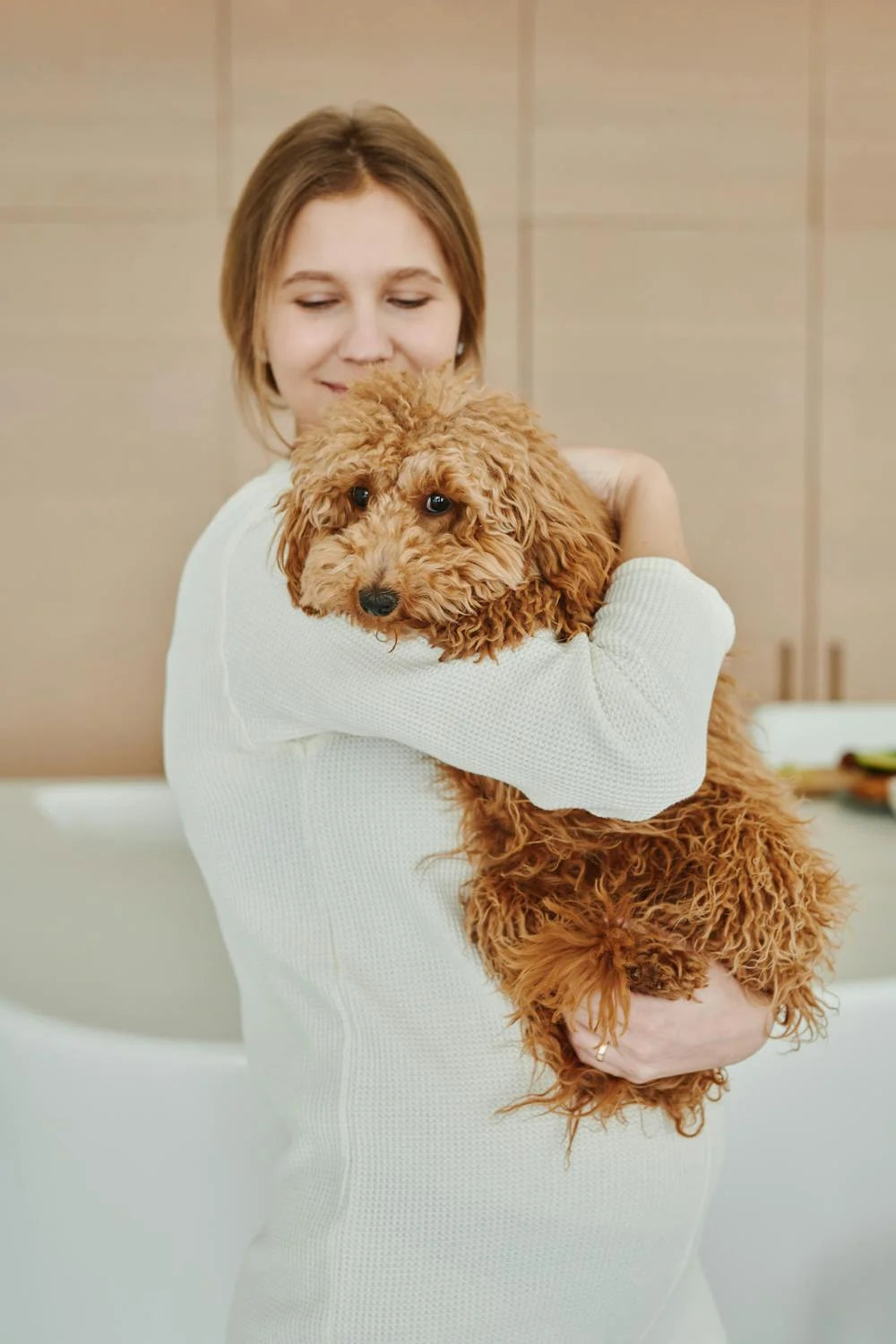 Woman holding shaggy golden doodle in a bathroom, near a bathtub. 